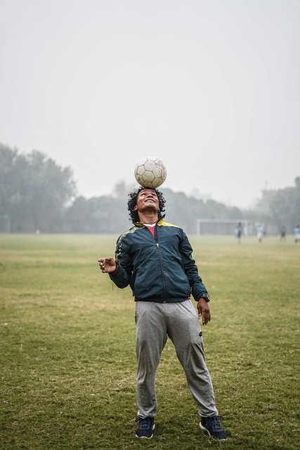 A man with black curly standing on a grass soccer feild with a white soccer ball balancing on his forhead along with a smile as big as a water melon and a blue jacket and gray sweat pants