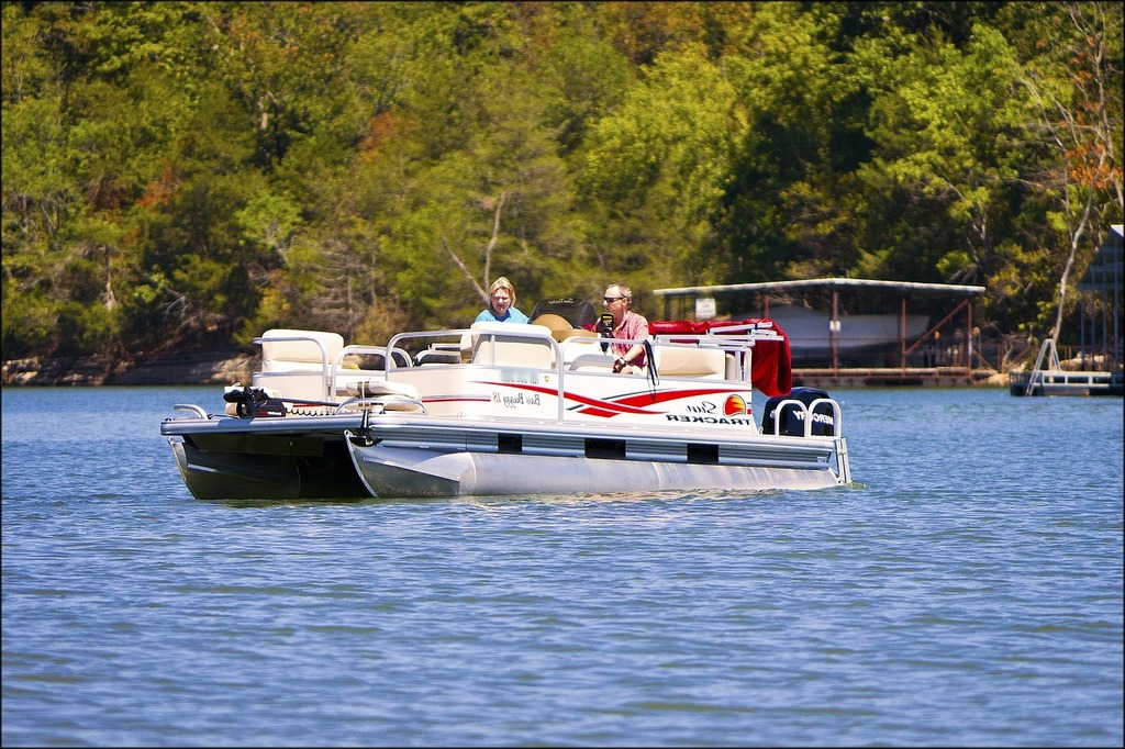 A pontoon boat on a lake with two people on it with trees in the background on the shore of the lake