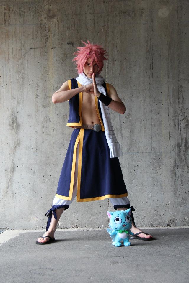 shirtless pink haird man with a yellow and black sleeveless jacket, white pants with a black and yellow covering, a white scarf, black sandals, and a blue cat plush