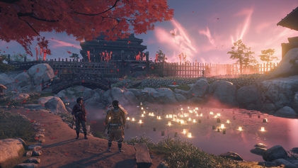 Jin Sakai and Yuna standing on a dirt path in front of a small, glistening lake under a bridge lit up by paper lanterns with an orange and rose sunset in the background